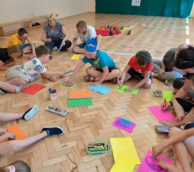 Dzieci i dorośli siedzą na podłodze, rysując i bawiąc się. Wszyscy są zaangażowani w zabawy integracyjne, tworząc kolorowe prace.