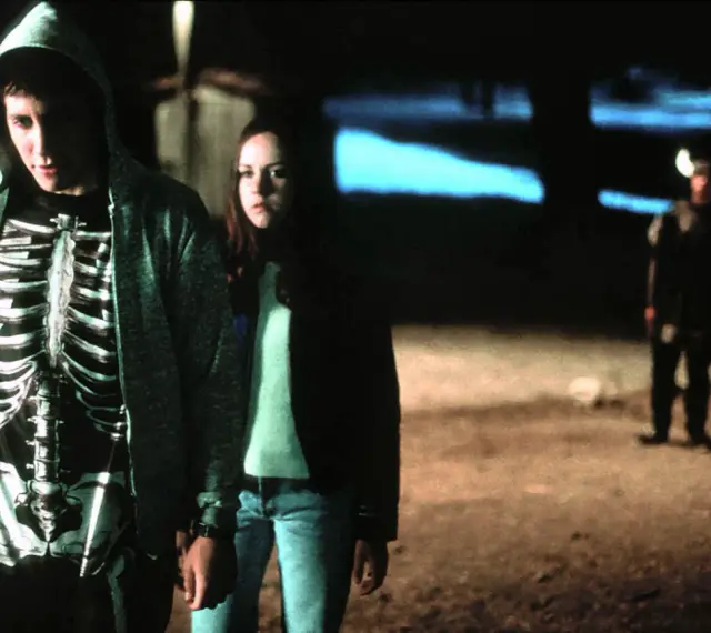 Donnie Darko w bluzie z kapturem i koszulce z nadrukiem szkieletu, obok dziewczyny. W tle dwie postacie w kostiumach.