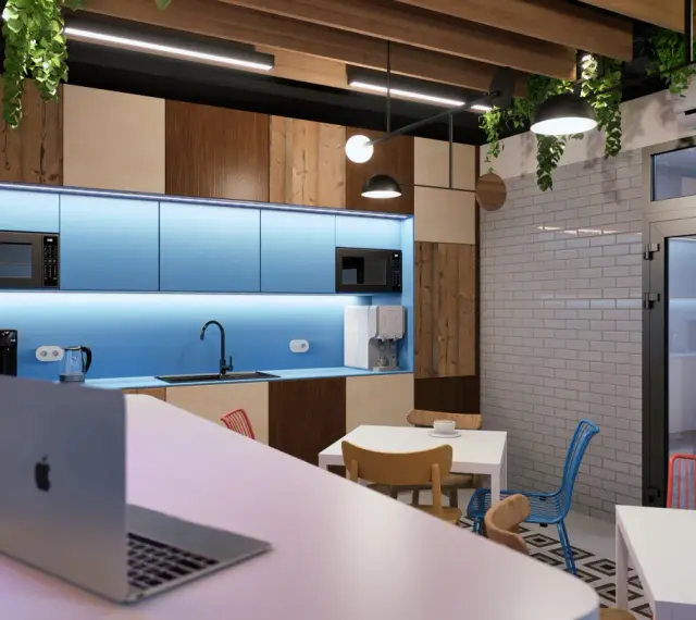 Nowoczesne pomieszczenie socjalne z niebieskimi szafkami, drewnianymi panelami, stołami i krzesłami. Laptop na blacie zaprasza do pracy.