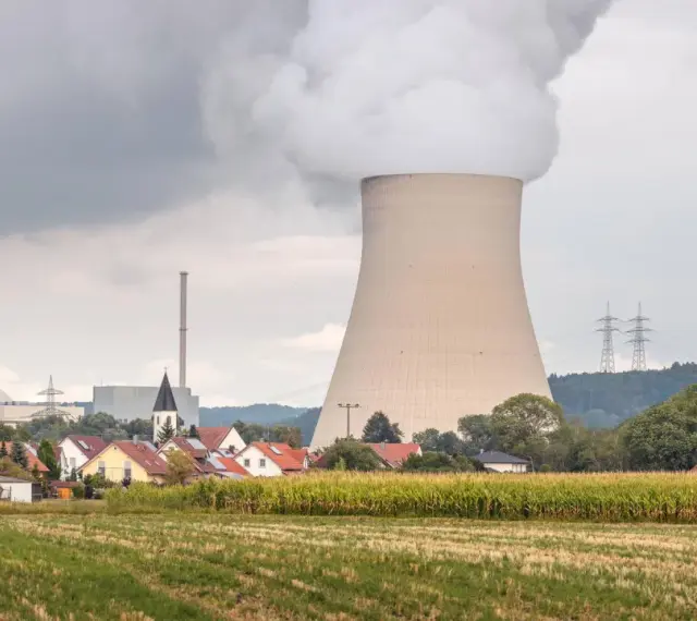 Dampf steigt aus einem Kühlturm eines Atomkraftwerks auf, daneben ein Dorf und ein Maisfeld.