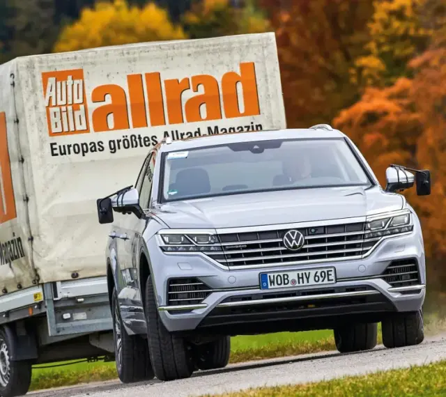 Srebrny Volkswagen Touareg holuje przyczepę z logo "Auto Bild allrad", jadąc po krętej drodze wśród jesiennych drzew.