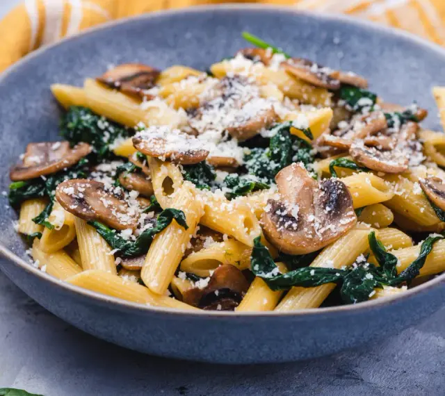 Penne z pieczarkami i szpinakiem, posypane parmezanem, w szarej misce.