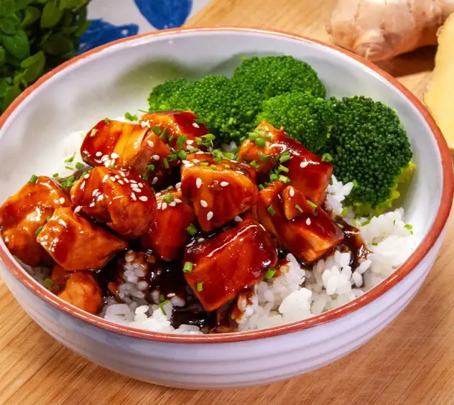 Miseczka ryżu z kawałkami lśniącego lososia teriyaki, posypanego sezamem i szczypiorkiem, obok parujących różyczek brokułów.