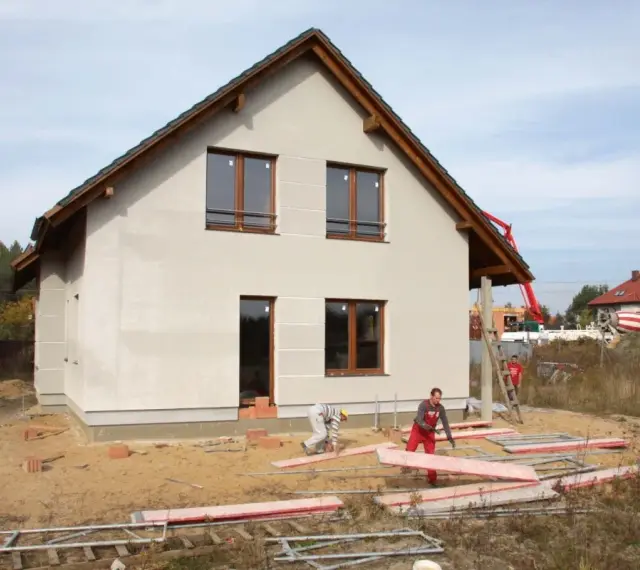 Budowa domu w toku. Pracownicy układają płyty, a w tle widać betonowóz. Czas budowy zależy od wielu czynników.