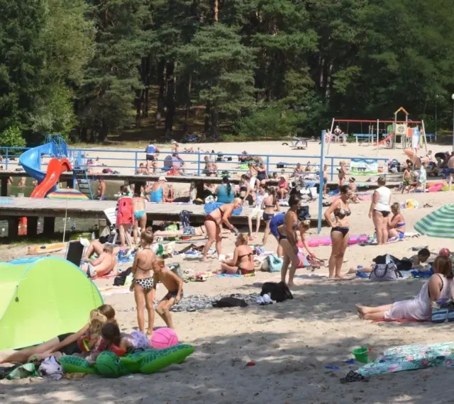 Tłoczna plaża nad jednym z **jezior blisko Wrocławia**. Ludzie opalają się, kąpią i odpoczywają na piasku.