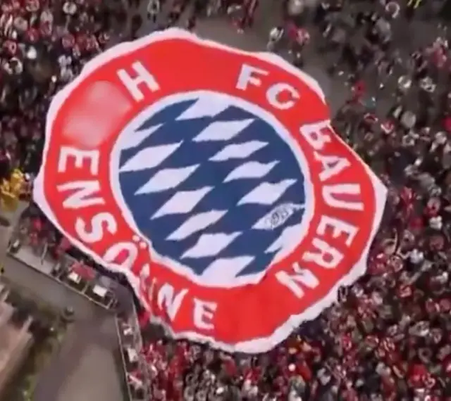 Ogromna flaga z herbem FC Bayern Monachium niesiona przez tłum kibiców.