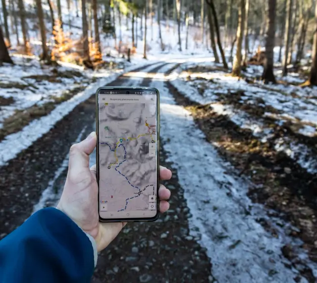 Telefon z mapą pokazuje szlak w lesie. Widać ślady śniegu na drodze, a na ekranie zaznaczony jest skopiec szlak.