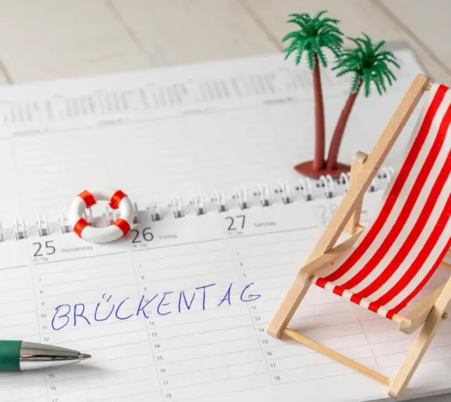 Kalenderblatt mit "Brückentag" markiert. Mini-Strandstuhl, Palmen und Rettungsring deuten auf Pfingstferien 2026 in Bayern hin.