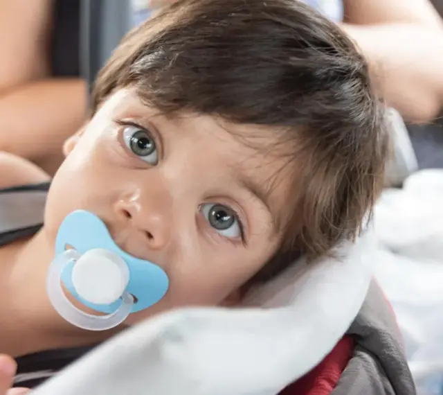 Małe dziecko z niebieskim smoczkiem Tommee Tippee w buzi, zapatrzone w dal.