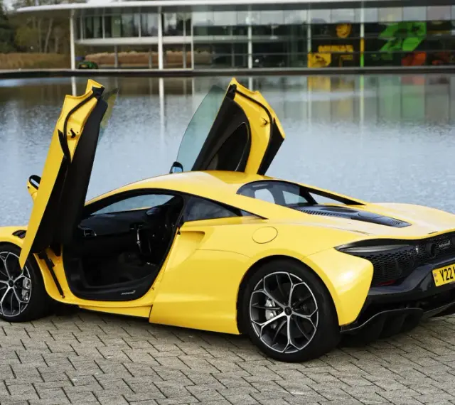 Żółty McLaren Artura z otwartymi drzwiami, stojący nad jeziorem przy nowoczesnym budynku.