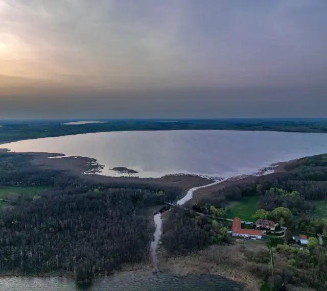 Rozległe jezioro Tuchlin otoczone lasem, z widocznym budynkiem i drogą. Zachód słońca maluje niebo.