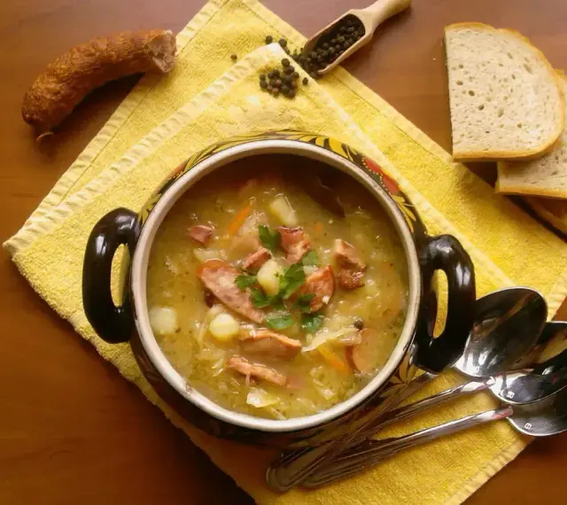 Aromaticzny kapuśniak z kiełbasą i ziemniakami, podany w ozdobnym naczyniu, obok kromek chleba i ziaren pieprzu.