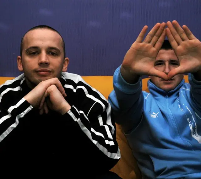 Dwóch mężczyzn w dresach, jeden z rękami złożonymi na brodzie, drugi tworzy rękami kształt serca. Klimat hip hop i rap.