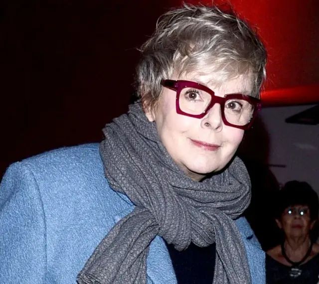 Karolina Korwin Piotrowska w okularach i niebieskim płaszczu, z szarym szalem na szyi.