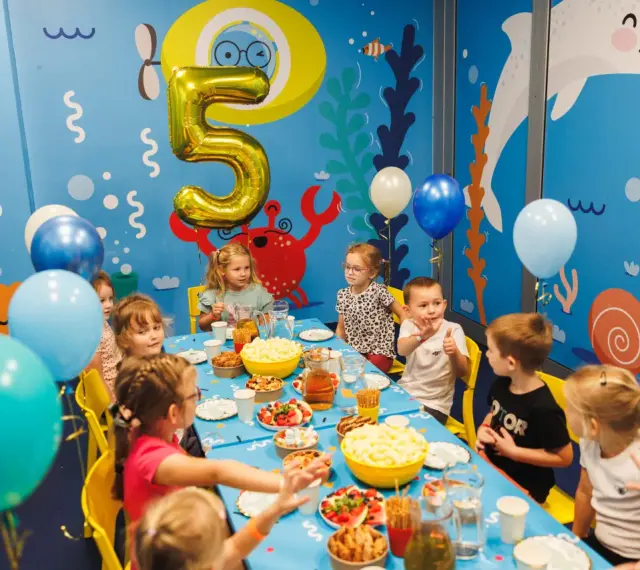 Dzieci świętują 5 urodziny przy stole pełnym przekąsek. Złoty balon w kształcie cyfry 5 unosi się nad stołem.