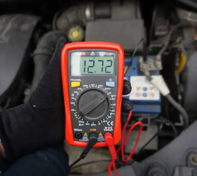 Ręka w czarnej rękawiczce trzyma czerwony multimetr z wyświetlaczem 12.72V, na tle silnika samochodu.