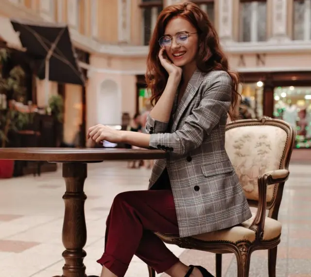 Elegancka kobieta w okularach i marynarce w kratę, prezentująca stylizacje z bordowymi spodniami, siedzi przy stoliku w kawiarni.