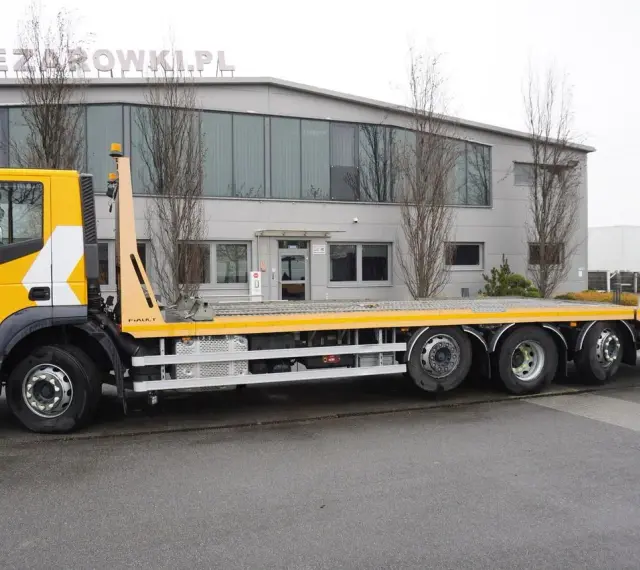 Żółty Iveco Stralis z platformą transportową stoi przed budynkiem.