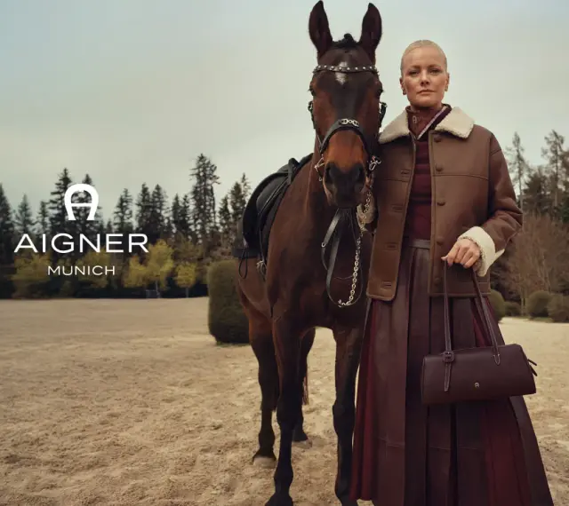 Elegancka kobieta w brązowej kurtce i spódnicy obok konia. Logo AIGNER MUNICH. Aigner co to za marka? Luksusowa moda.