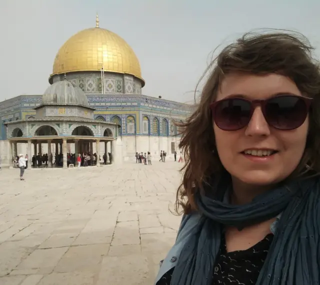Uśmiechnięta kobieta w okularach przeciwsłonecznych robi selfie na tle Kopuły na Skale. Wokół widać turystów, co sugeruje, że w Jerozolimie jest bezpiecznie.