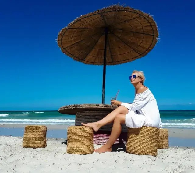 Kobieta w białej sukience relaksuje się pod parasolem na plaży, popijając napój. Idealne tunezja wakacje.