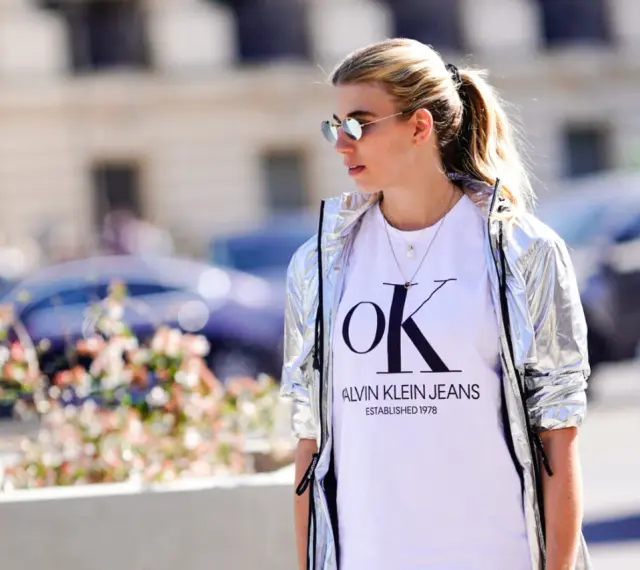 Kobieta w srebrnej kurtce CK damskiej i białej koszulce z logo Calvin Klein Jeans.