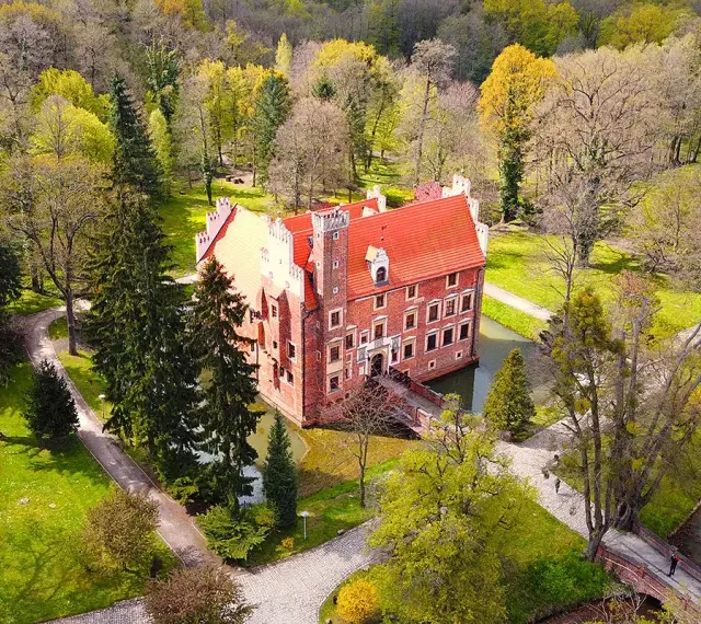 Zamek na wodzie w Wojnowicach otoczony zielenią i drzewami. Czerwona bryła zamku z charakterystycznym dachem wznosi się nad fosą.