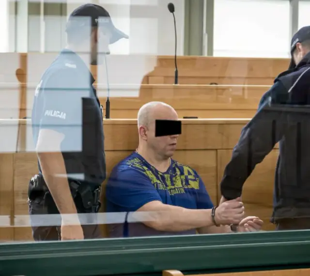 Skruszony gangster w kajdankach, prowadzony przez policjantów w sali sądowej.