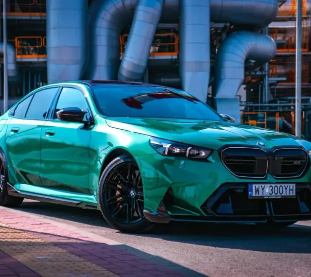 Błyszczące, zielone BMW M5 G90 z czarnymi felgami stoi na tle przemysłowych rur.