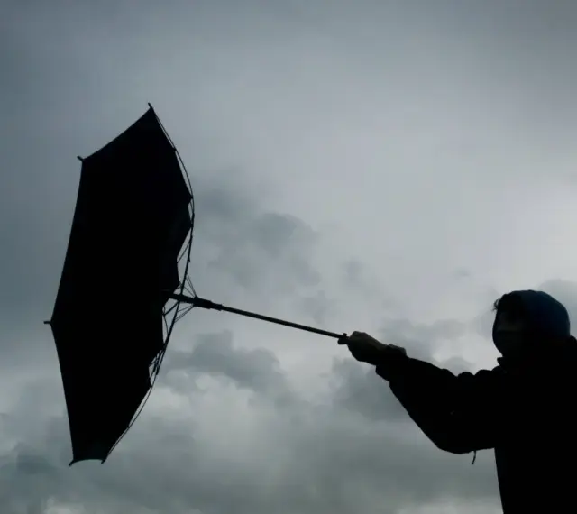 Silhouette einer Person mit einem aufgewehten Regenschirm vor dramatischem Himmel. Der **Orkan** kämpft gegen den Schirm.