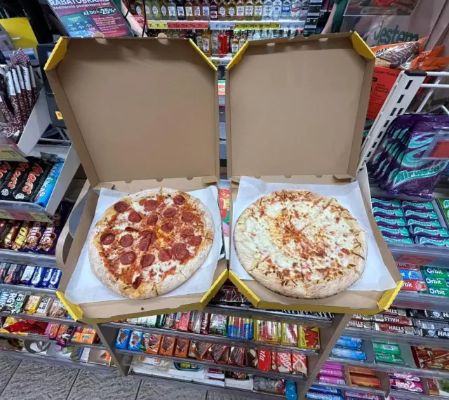 Dwie pizze w pudełkach, jedna z salami, druga serowa. Obok półki ze słodyczami i napojami w sklepie Żabka.