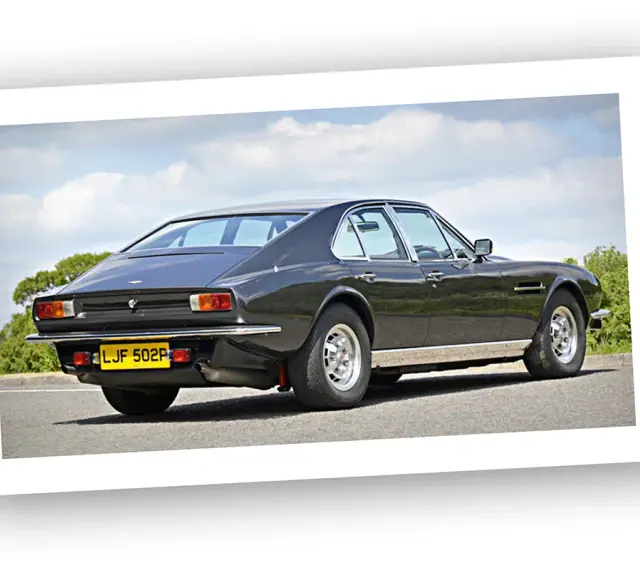 Klasyczny Aston Martin Lagonda w ciemnym kolorze, widziany z tyłu, na tle błękitnego nieba z chmurami.