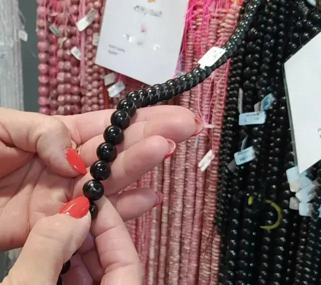 Manos con uñas rojas sostienen un collar de cuentas negras de shungita. En el fondo, se ven hilos de cuentas rosas y blancas.