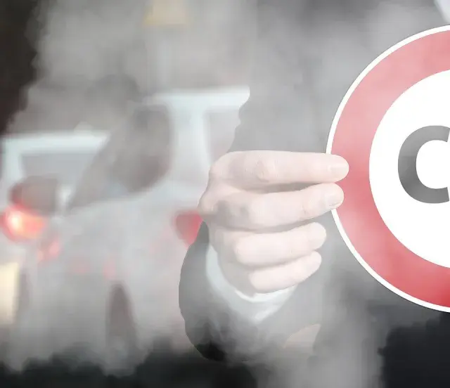 Dłoń trzyma znak zakazu z napisem CO2 na tle dymiących samochodów w korku.