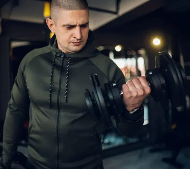 Mężczyzna w zielonej bluzie damskiej na siłownię ćwiczy biceps z hantlem.
