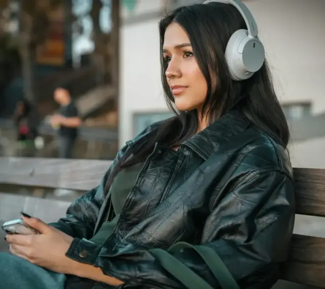 Młoda kobieta w słuchawkach, słuchająca podcastu o wymowie francuskiej, siedzi na ławce, trzymając telefon.
