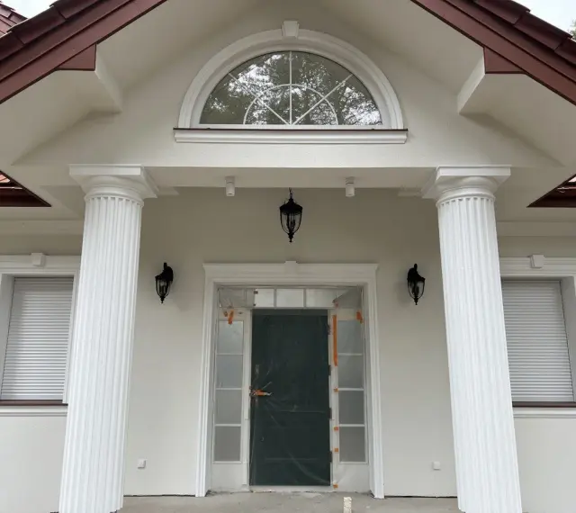 Elegancki pilaster zdobi fasadę domu, wspierając dach nad wejściem. Obok okna i drzwi, tworzy klasyczną kompozycję.