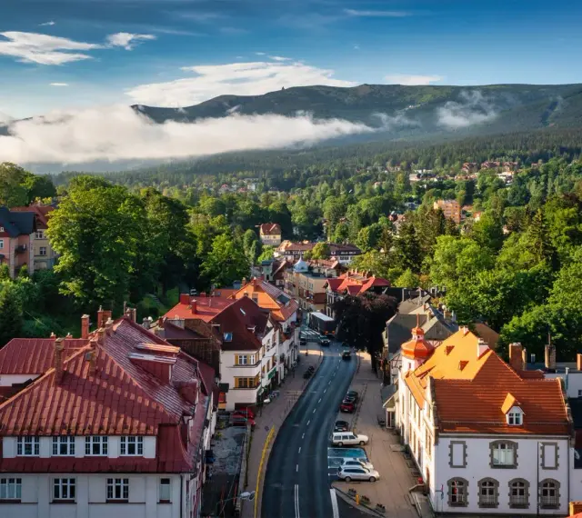 Widok z góry na miasteczko z czerwonymi dachami, otoczone zielonymi lasami i górami w tle.