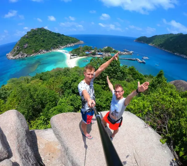 Dwóch mężczyzn robi selfie na tle tropikalnej wyspy z turkusową wodą i plażą.