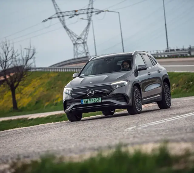 Szary Mercedes-Benz EQA jedzie po drodze, w tle linie energetyczne i drzewo.