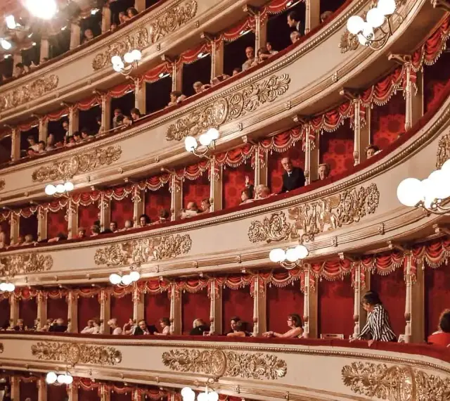 Widownia La Scali, pełna widzów w lożach, oczekuje na rozpoczęcie spektaklu. Bogaty repertuar teatru przyciąga miłośników sztuki.