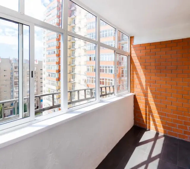 Wnętrze loggii z widokiem na miejską zabudowę. Ściany z cegły i tynku, przeszklony balkon z białymi ramami okiennymi.