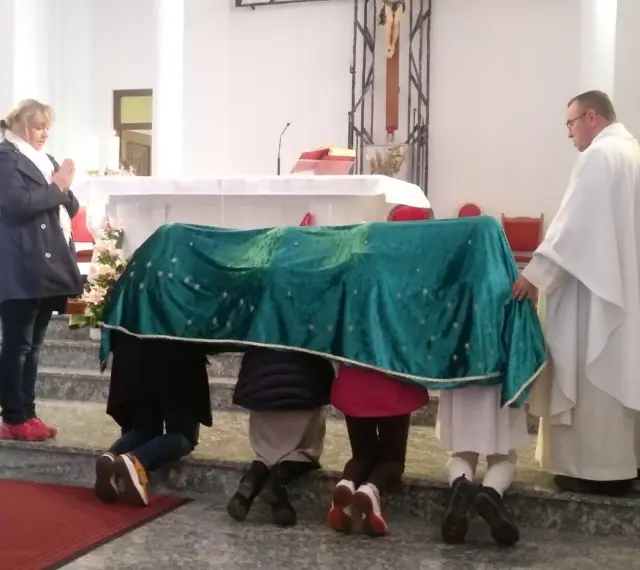 Dzieci klęczą pod zielonym welonem, odmawiając modlitwę do Matki Bożej z Guadalupe. Kapłan czuwa obok.