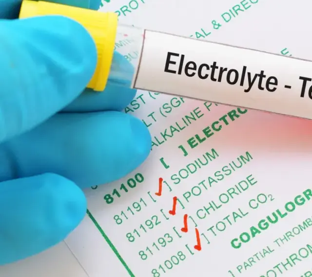 Ręka w niebieskiej rękawiczce trzyma probówkę z oznaczeniem "Electrolyte - Test" nad formularzem z badaniami, w tym jonogramem.
