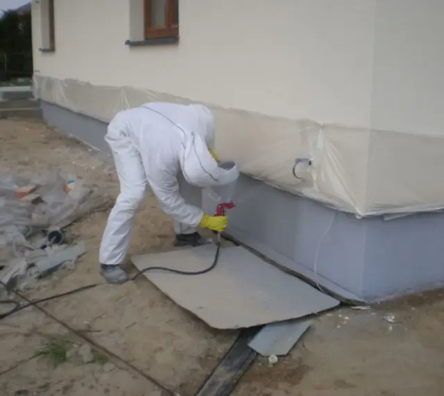 Pracownik w białym kombinezonie maluje fundamenty na kolor przypominający rubinowy.