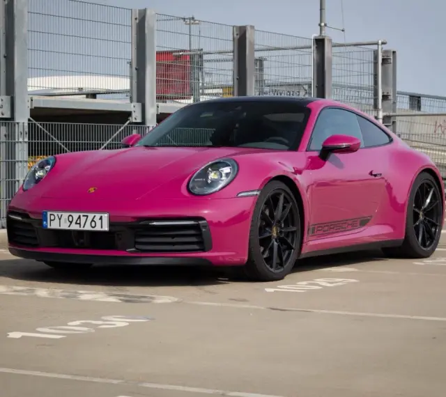 Różowy Porsche 911 Carrera 4S z czarnymi felgami i naklejką PORSCHE na progu, zaparkowany na zewnątrz.