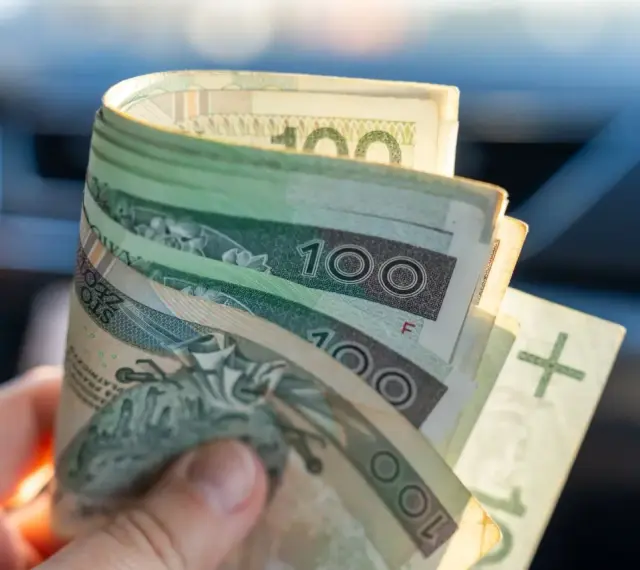 Dłoń trzyma zwitek polskich banknotów 100 zł. Dokument orzeczenie o niepełnosprawności może pomóc w uzyskaniu zasiłek pielęgnacyjny.