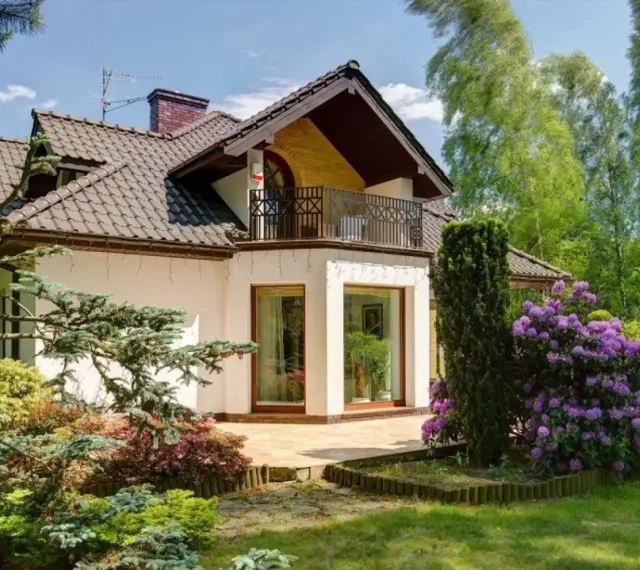 Dom z balkonem i tarasem, otoczony bujną zielenią i kwitnącymi rododendronami. Występ ściany podkreśla nowoczesny charakter budynku.