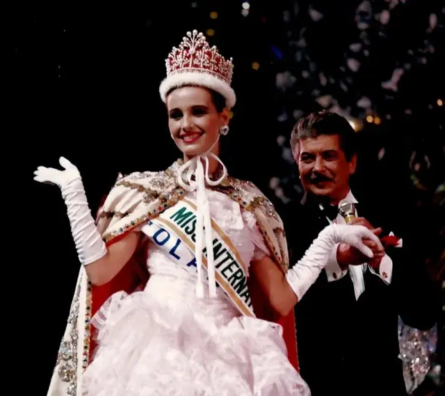 Uśmiechnięta Miss International w koronie i białej sukni, z szarfą "Miss Polonia". Obok niej mężczyzna w smokingu.