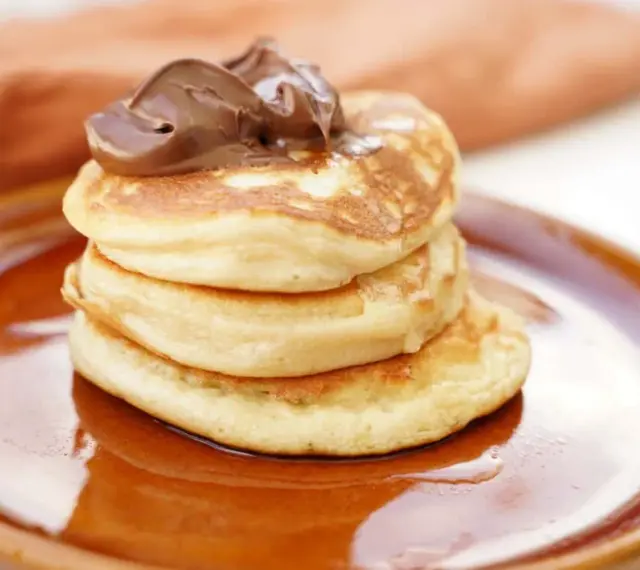 Stack of three fluffy American pancakes topped with chocolate spread on a brown plate.